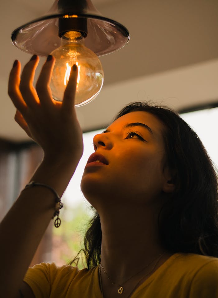 Home A young woman gazes thoughtfully at a glowing light bulb indoors, capturing a serene moment.